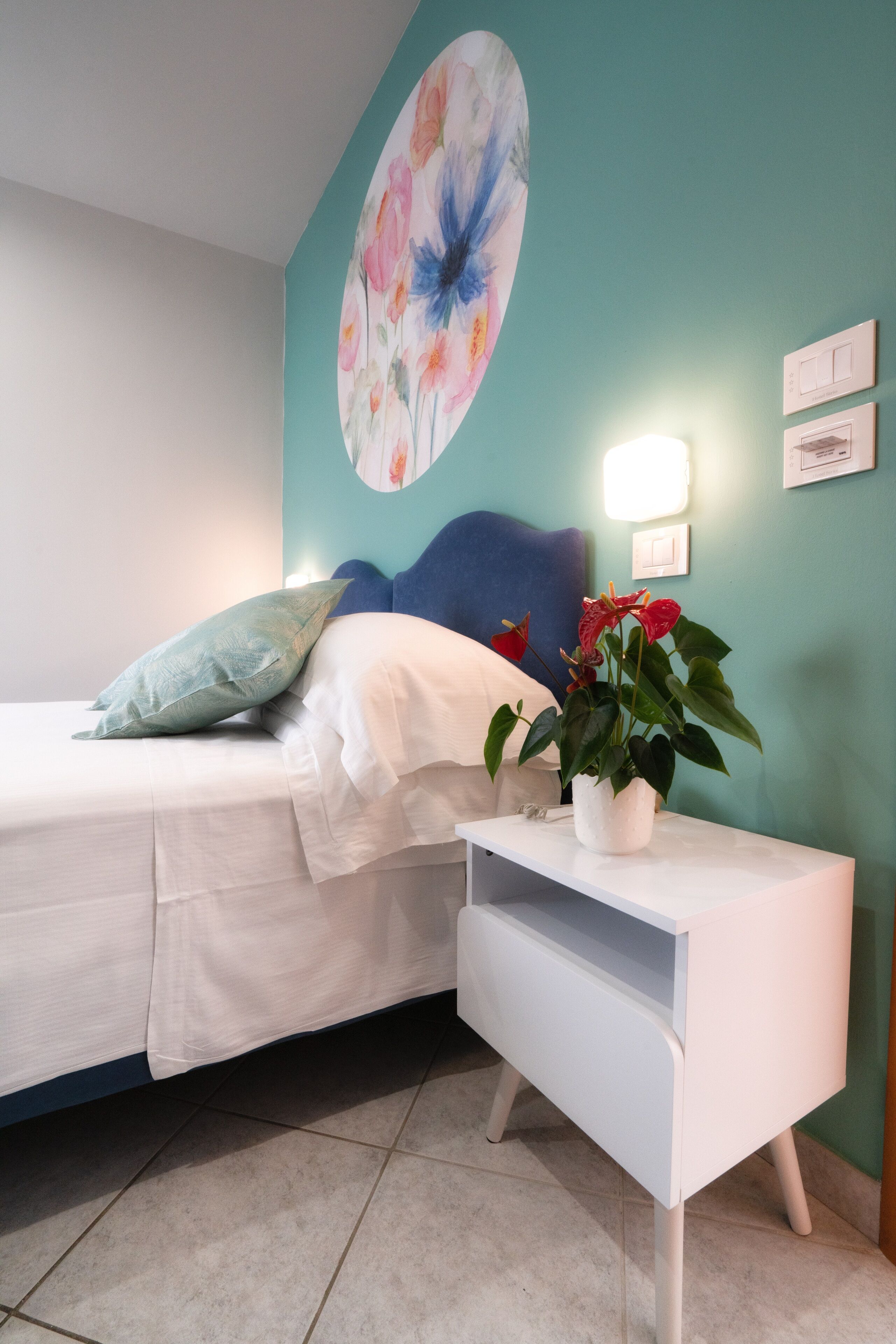 Ein Schlafzimmer mit einem Bett, einem Nachttisch mit Pflanze, einer Wand mit Blumenbild und einem blauen Bettvorhang.