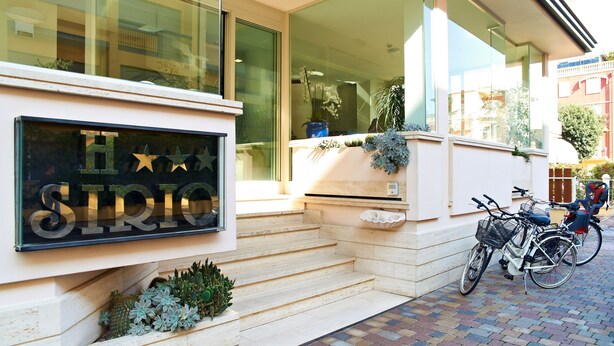 A building entrance with a sign reading "SIRIC," steps leading up to the door, and two bicycles parked outside. - Hotel Sirio (Camaiore)