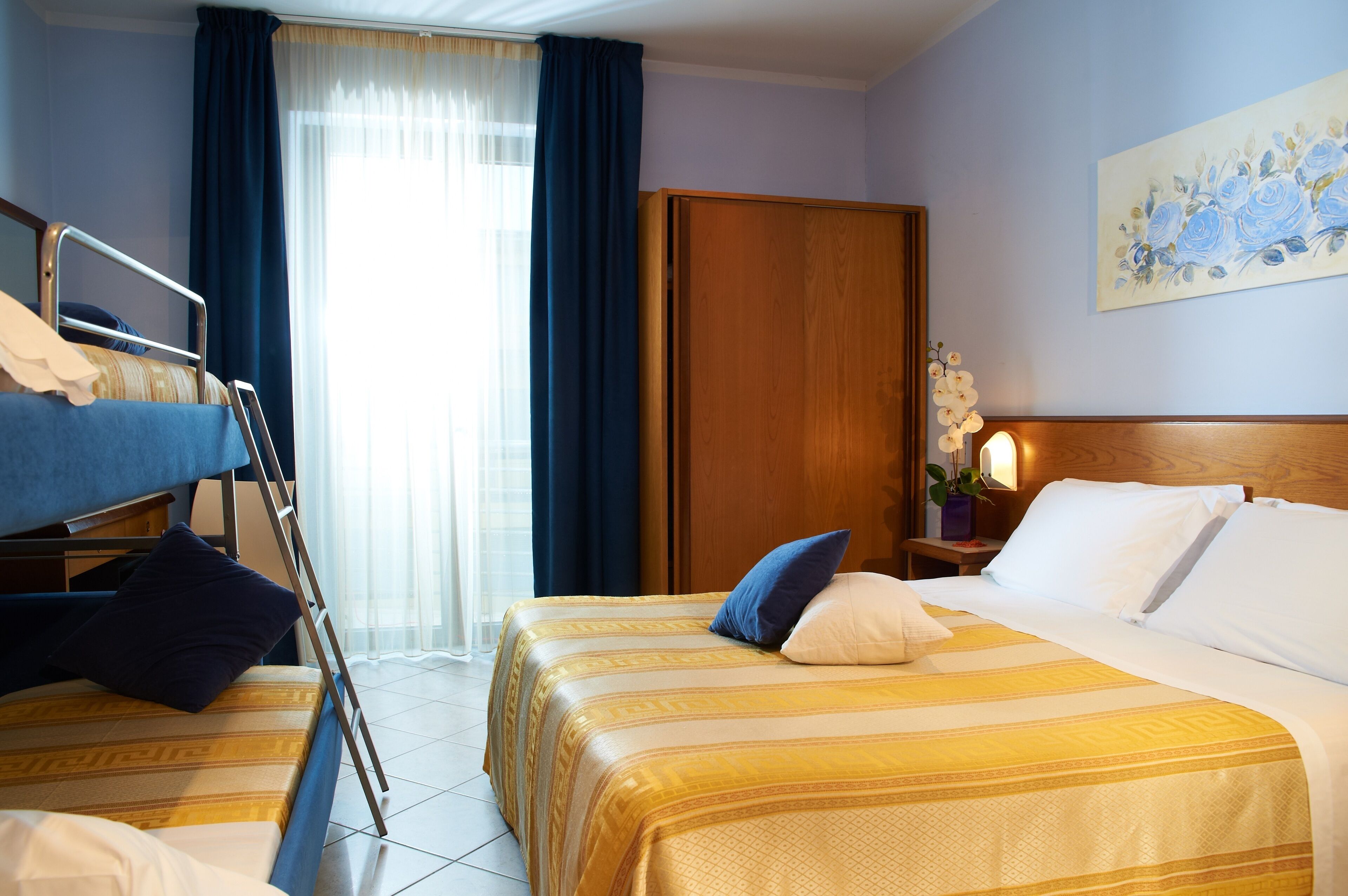 A hotel room with two beds, a bunk bed, a wooden wardrobe, a painting on the wall, and a nightstand with a lamp.