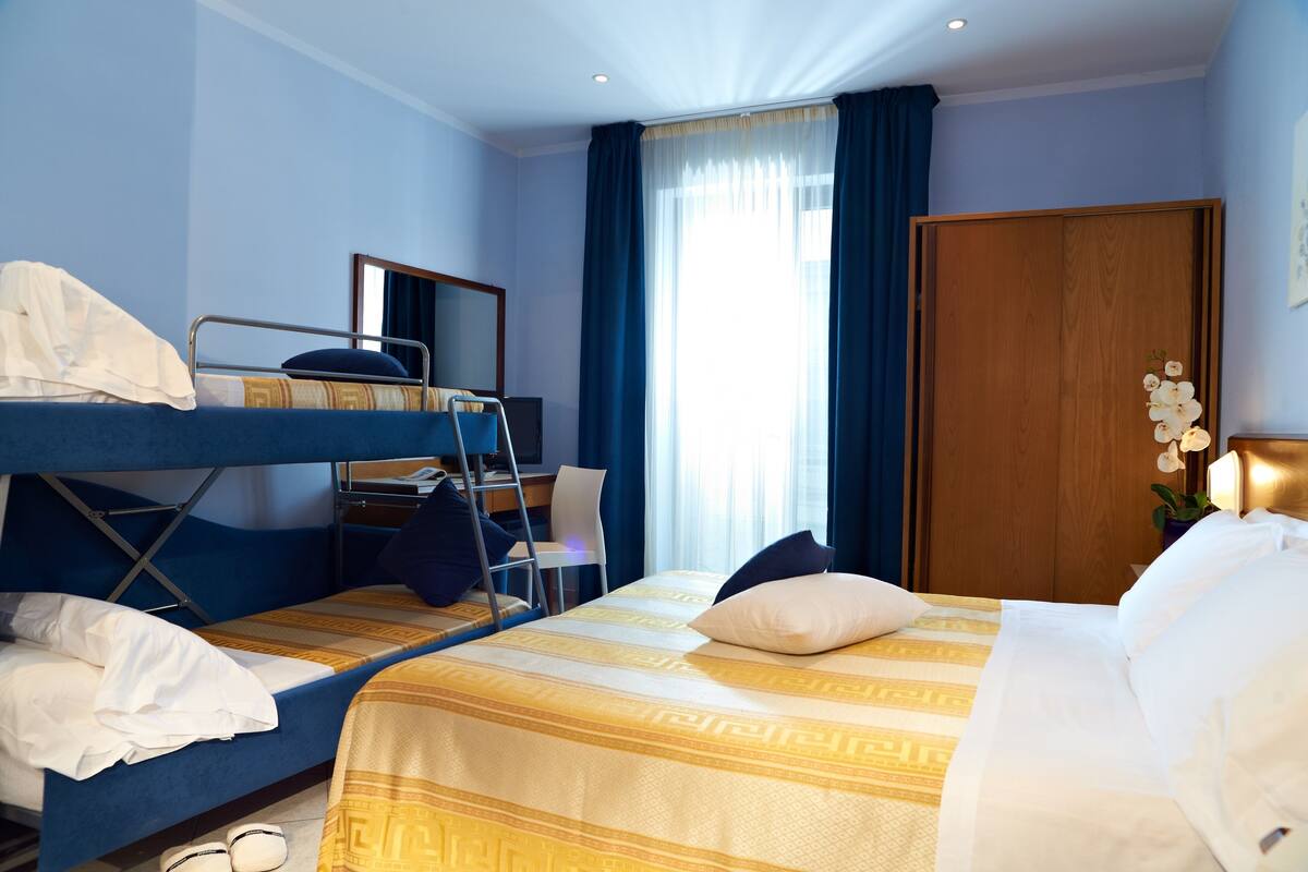 A hotel room with a bunk bed, a desk, a chair, a wardrobe, and a window with curtains.