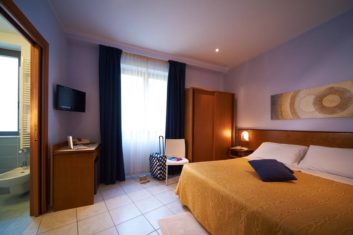 A hotel room with a bed, a desk, a chair, a television, a window with curtains, and a painting on the wall.
