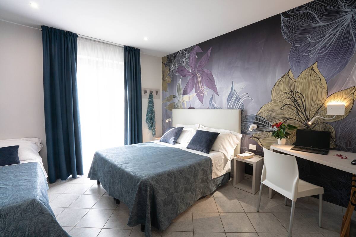 A hotel room with a bed, a desk with a laptop, a chair, and a wall with a floral mural.