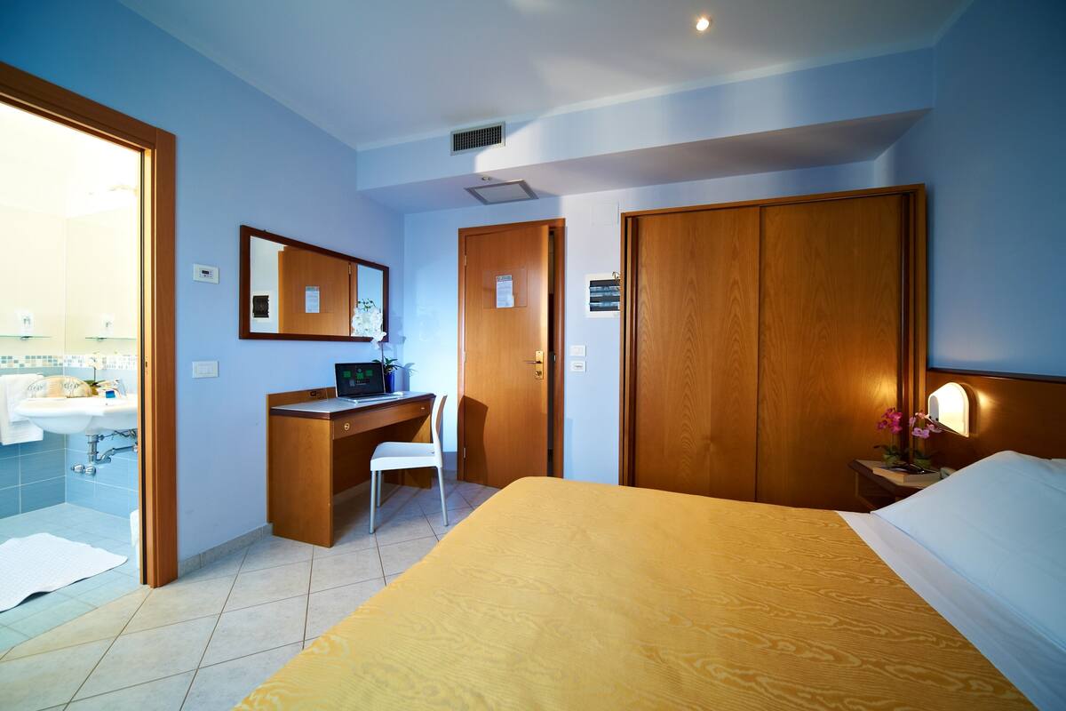 A hotel room with a bed, a desk, a chair, a bathroom with a sink, and a mirror.