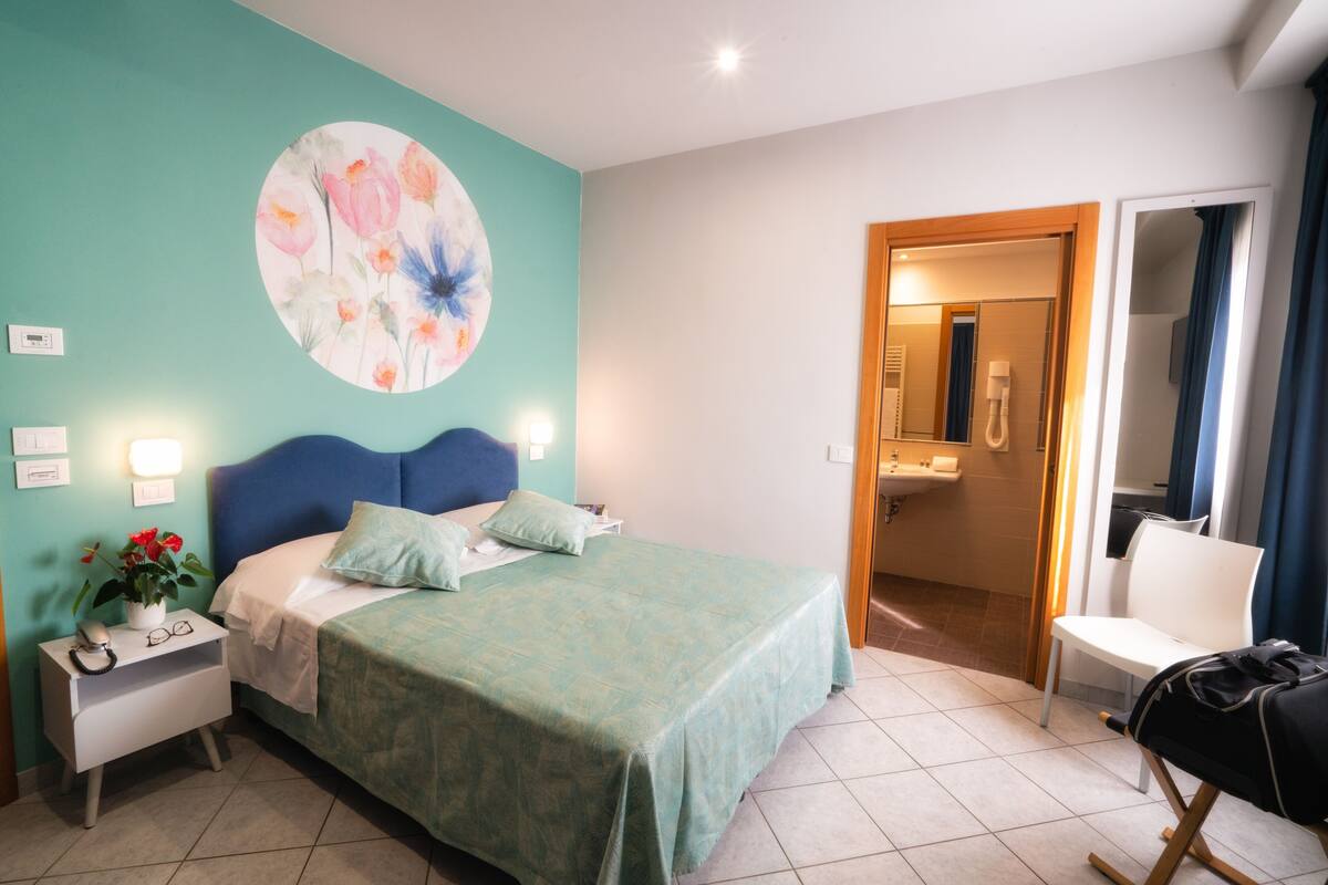 A bedroom with a bed, a nightstand, a wall painting, a bathroom visible through an open door, and a chair.