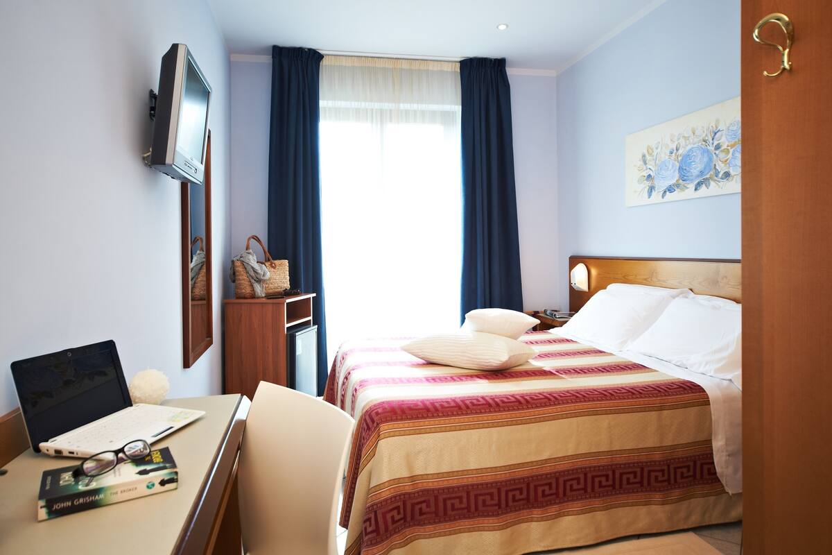 A hotel room with a bed, a desk with a laptop, a TV, and a window with curtains.