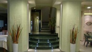 A hallway with a staircase leading to another room, potted plants on either side, and a chair in the corner. - Hotel Sirio (Camaiore)