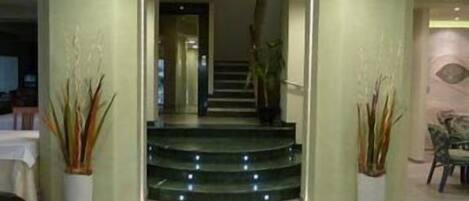 A hallway with a staircase leading to another room, potted plants on either side, and a chair in the corner.