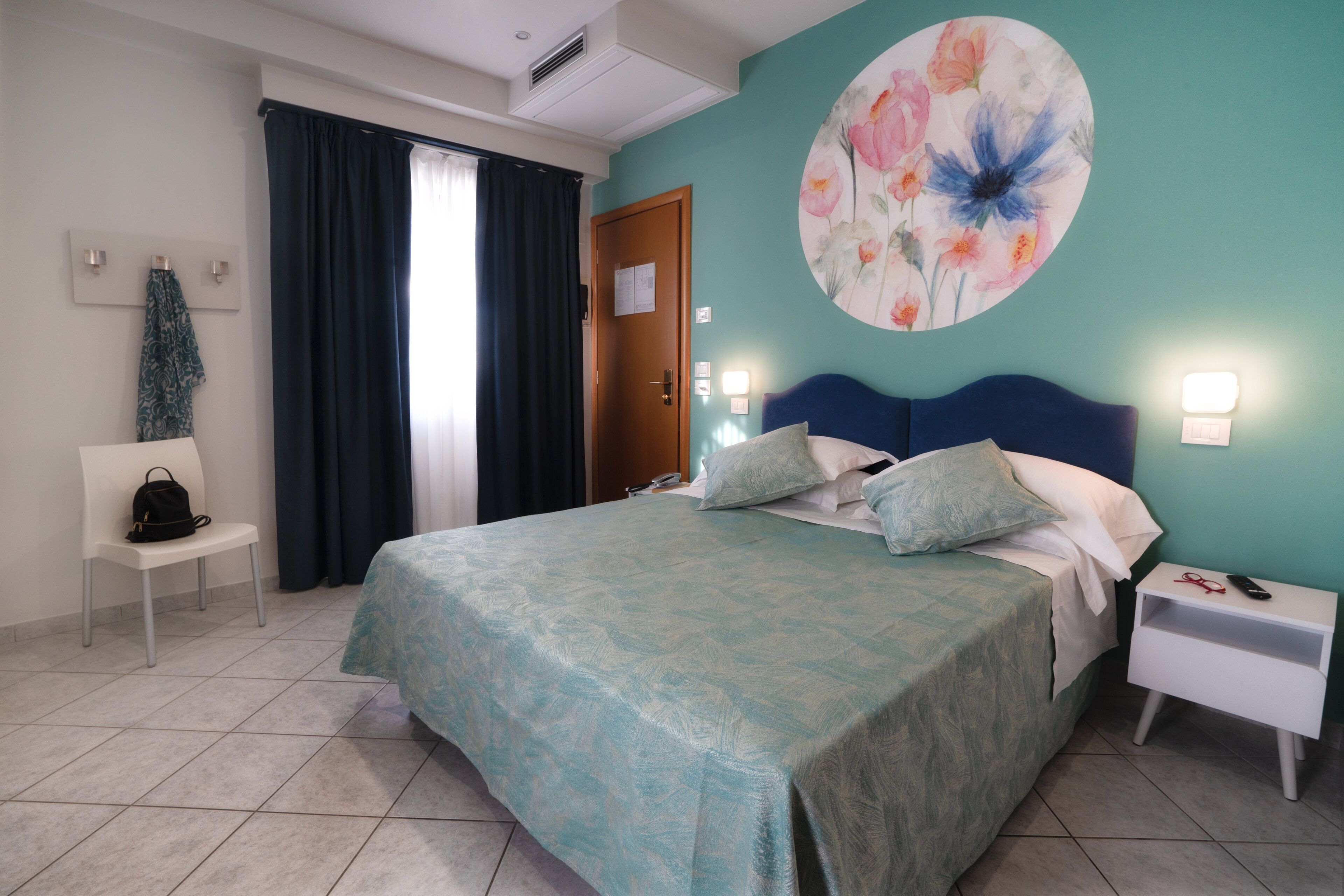 A bedroom with a bed, a nightstand, a chair, a door, and a wall with a floral painting.