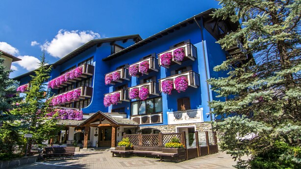 A blue building with balconies adorned with purple flowers, a wooden bench, and a stone entrance. - Hotel Baita Clementi (Bormio)