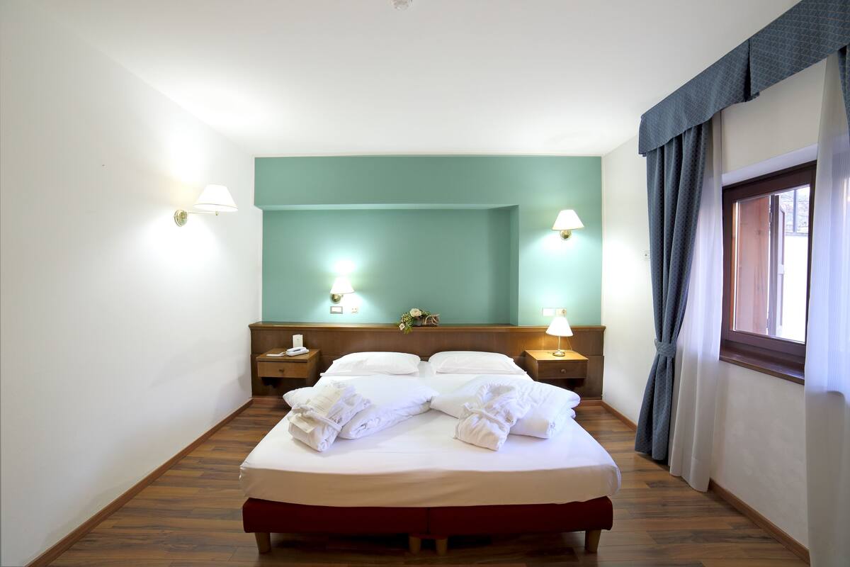 Ein ordentlich bezogenes Bett mit weißen Leinen und Handtüchern, zwei Nachttische mit Lampen, ein Holz-Kopfteil und ein Fenster mit Vorhängen.