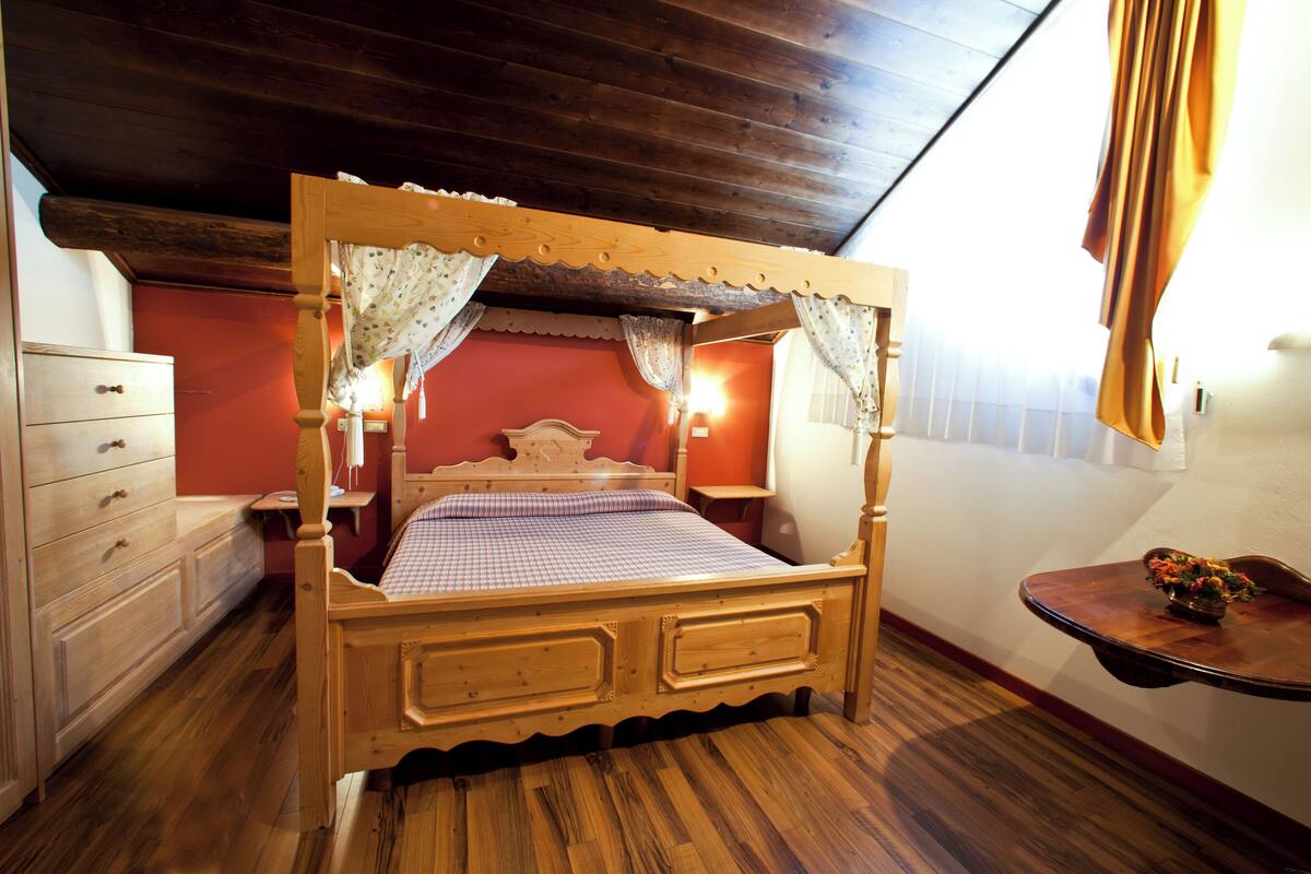 Ein hölzernes Himmelbett mit kariertem Überwurf und Spitzenvorhängen, eine Holzkommode und ein kleiner Tisch mit einer Vase.