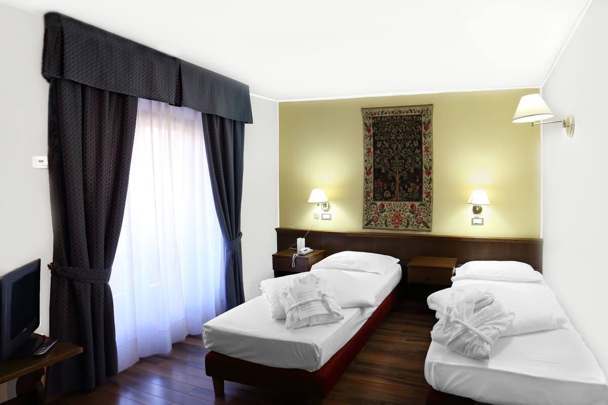 Ein Hotelzimmer mit zwei Einzelbetten, einem Fernseher, einem Schreibtisch und einem großen Fenster mit Vorhängen.