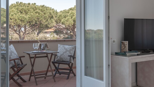 A balcony with a table, chairs, a TV, and a view of trees and houses. - Hotel President (Forte dei Marmi)