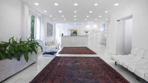 A spacious living room with a white sofa, a large red rug, and a potted plant. - Hotel President (Forte dei Marmi)