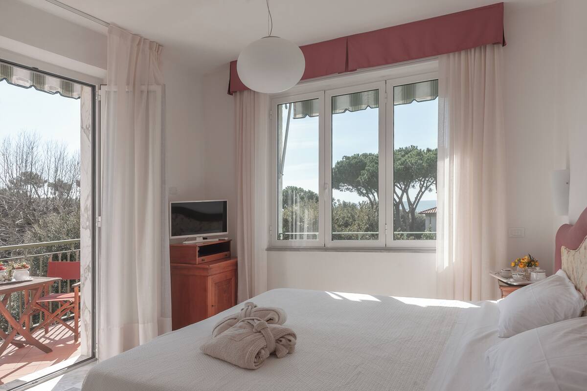 A bedroom with a bed, a TV, a balcony with a table and chairs, and a window with curtains.