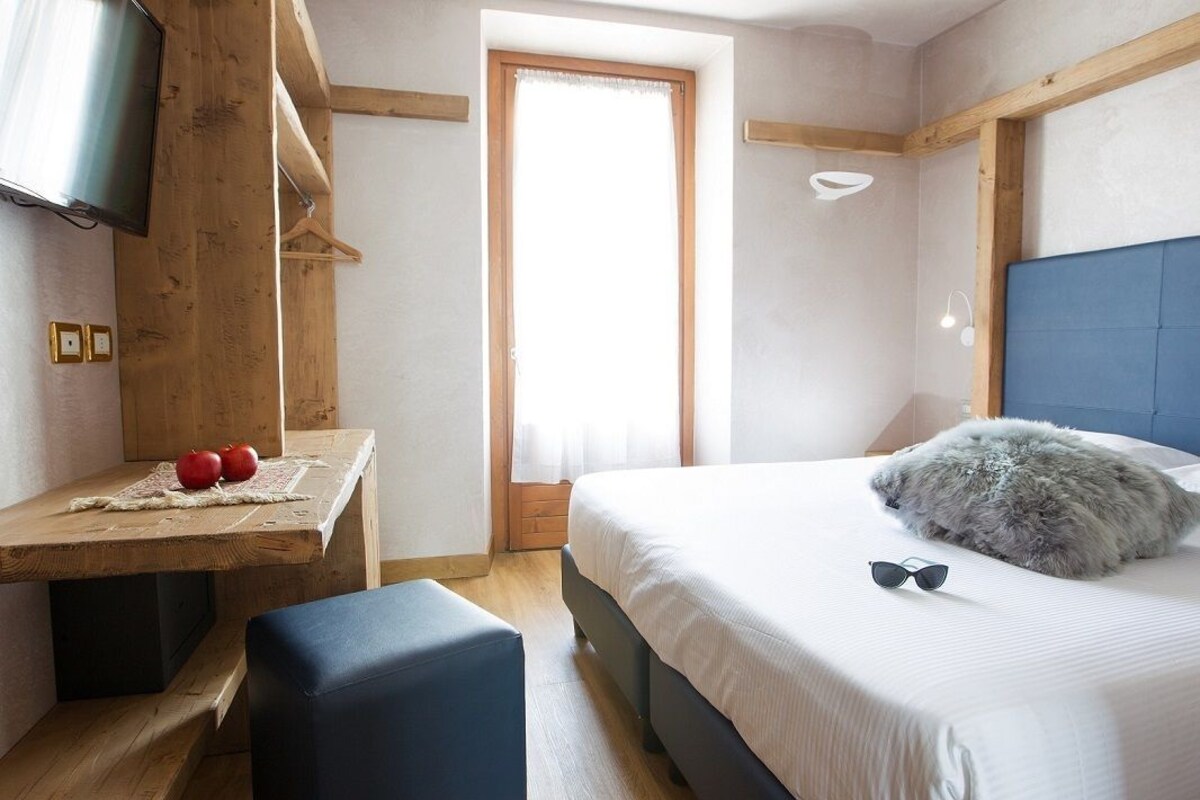 Un dormitorio con una cama, un televisor montado en una pared de madera, una mesita con manzanas y una ventana con cortinas.