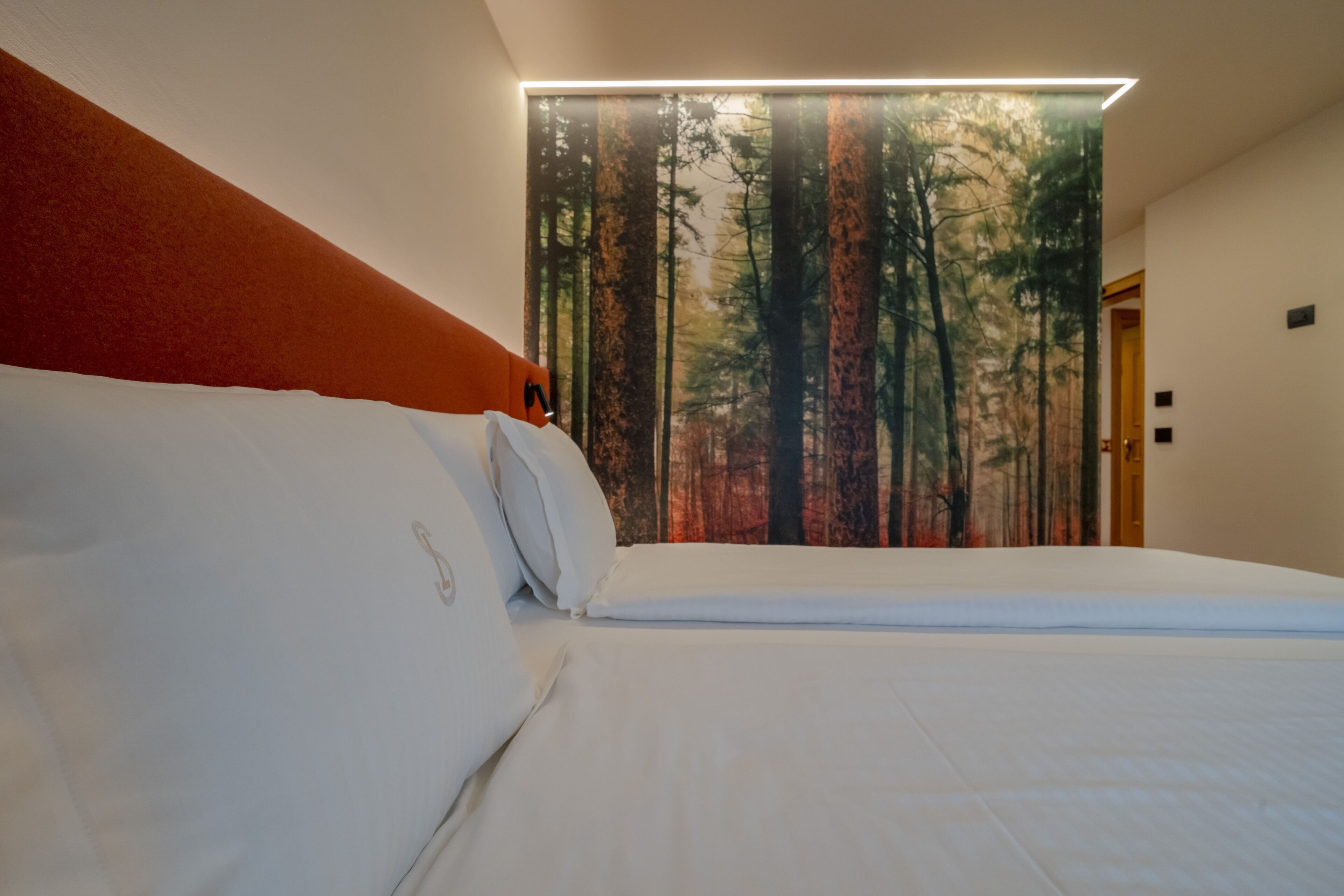 A neatly made bed with white linens and a headboard in a room with a forest-themed wall mural.