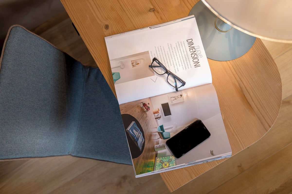 A magazine open on a wooden table, featuring a room layout with a bed, a chair, a lamp, and a smartphone.