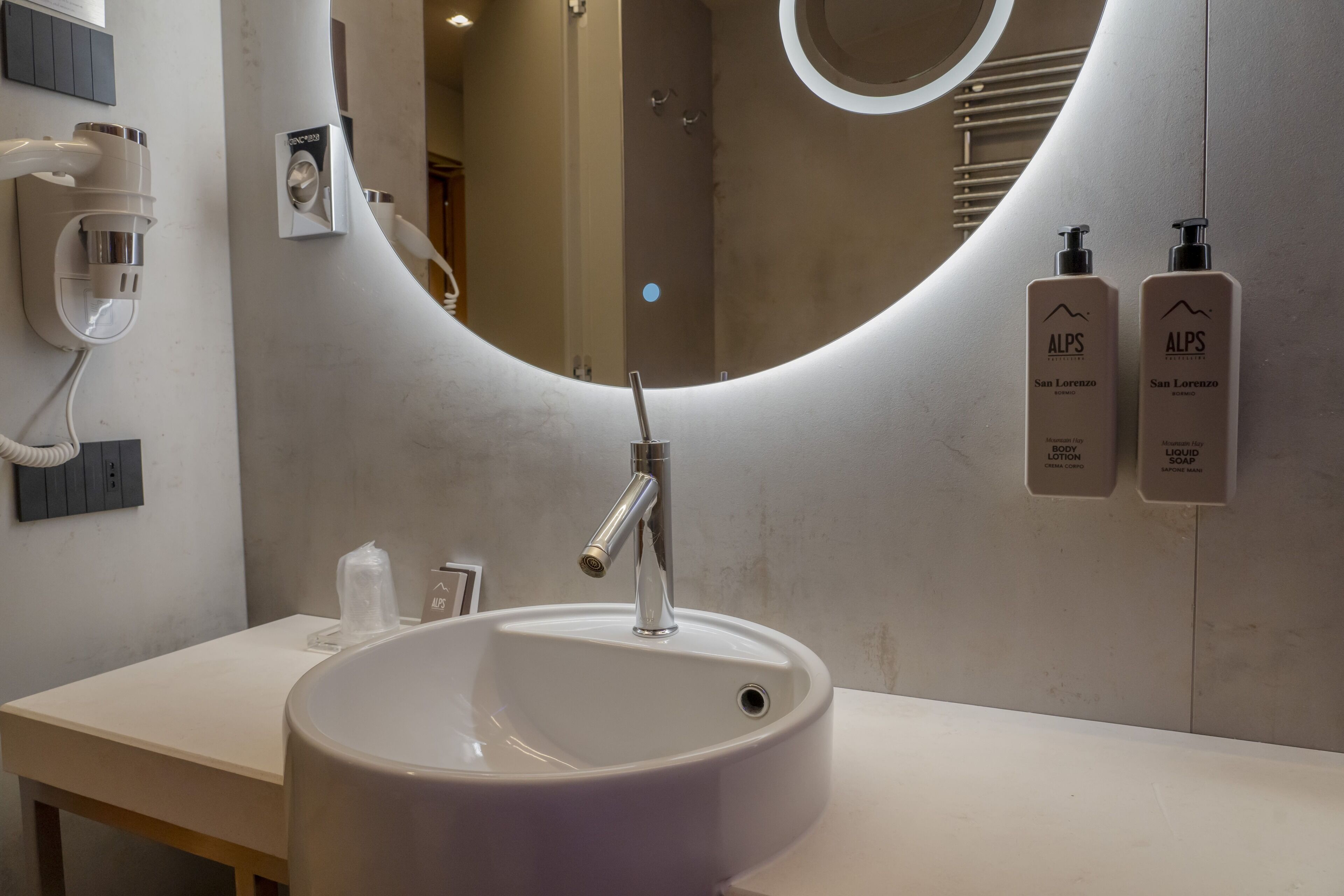 A modern bathroom with a large sink, a mirror, and two bottles of ALPS San Lorenzo products on the wall.