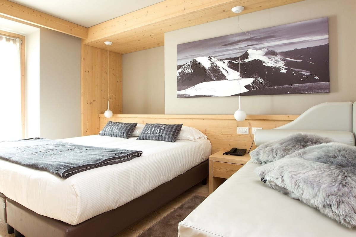 A modern bedroom with a wooden ceiling, a large bed with white bedding, a nightstand with a phone, and a mountain landscape painting on the wall.