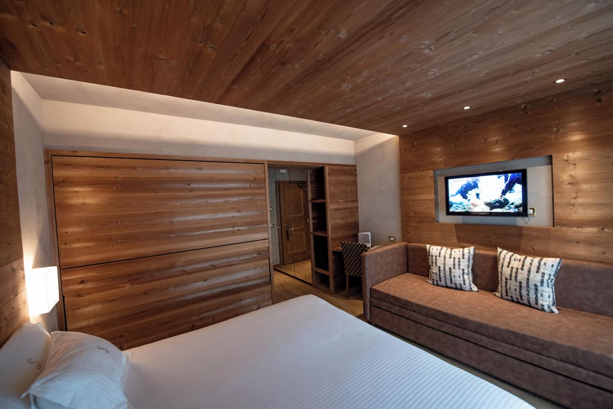 A modern bedroom with a wooden ceiling, a large bed, a sofa with patterned cushions, and a TV mounted on the wall.