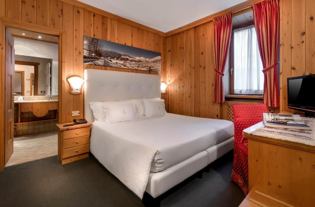 Une chambre à coucher aux murs lambrissés, avec un grand lit, une table de chevet, un miroir et un tableau représentant un paysage de montagne.