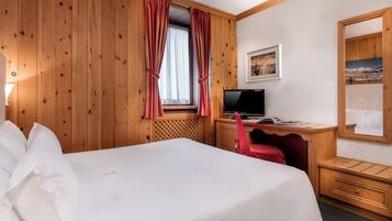 Habitación de hotel con pared de paneles de madera, cama con ropa de cama blanca, escritorio con computadora y ventana con cortinas rojas.