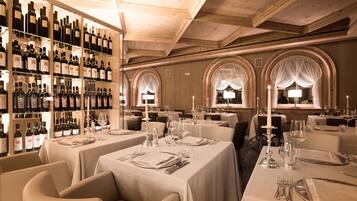 Un restaurant avec de nombreuses tables dressées pour le service, un présentoir à vin contenant différentes bouteilles et des fenêtres en arc surmontées d’un éclairage décoratif.