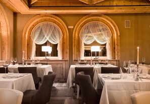 Un restaurante con varias mesas dispuestas para comer, con ventanales de arco y cortinas decorativas, y una iluminación cálida.