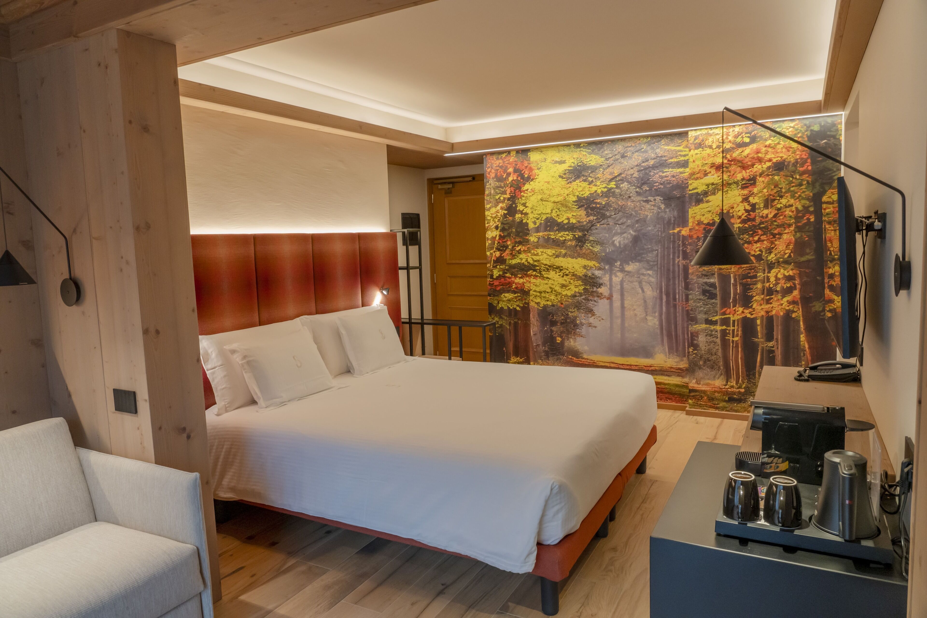 Une chambre moderne avec un grand lit, un bureau doté d’une machine à café et un décor mural représentant des arbres d’automne.