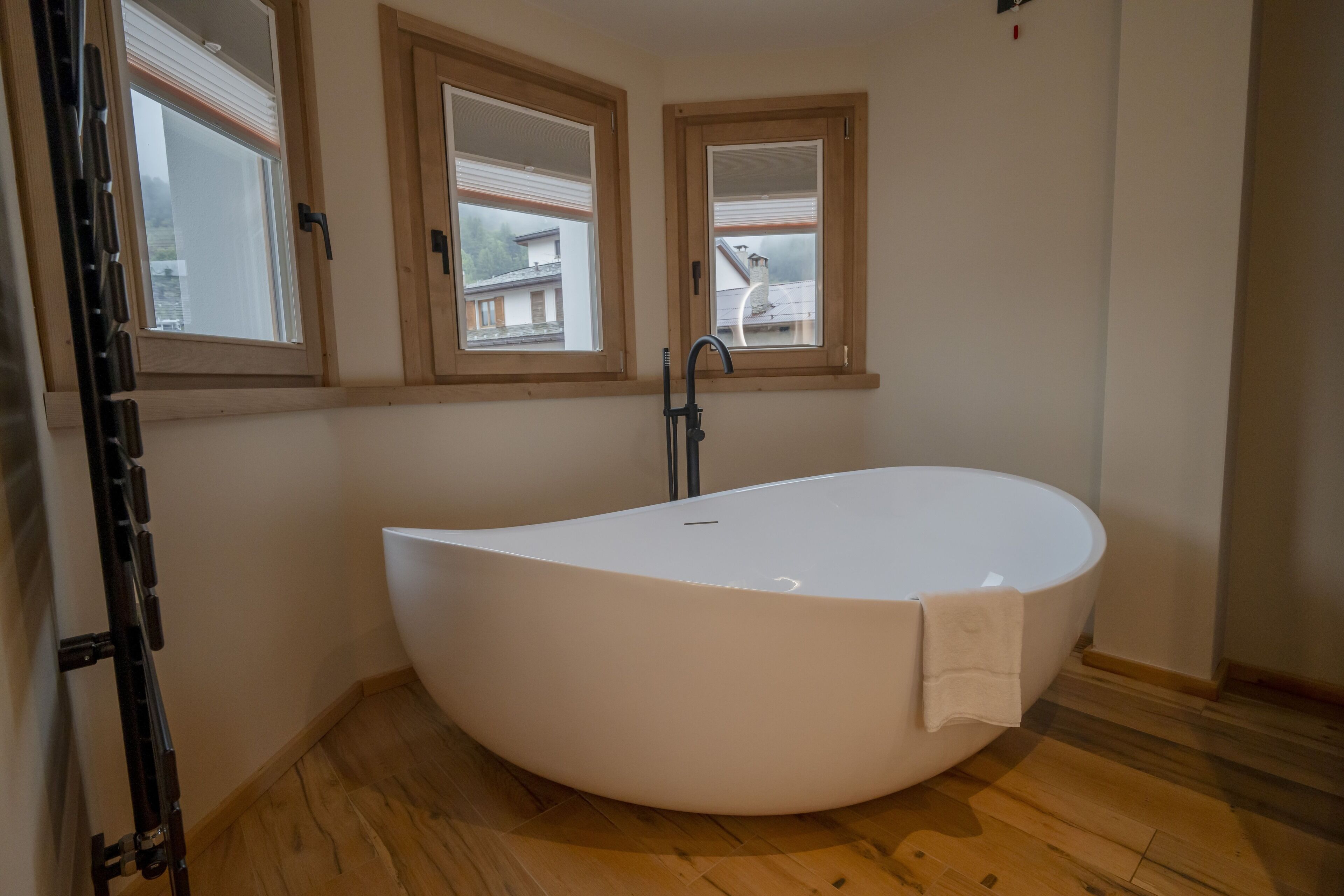 A modern bathroom with a freestanding bathtub, wooden flooring, and three windows with blinds.