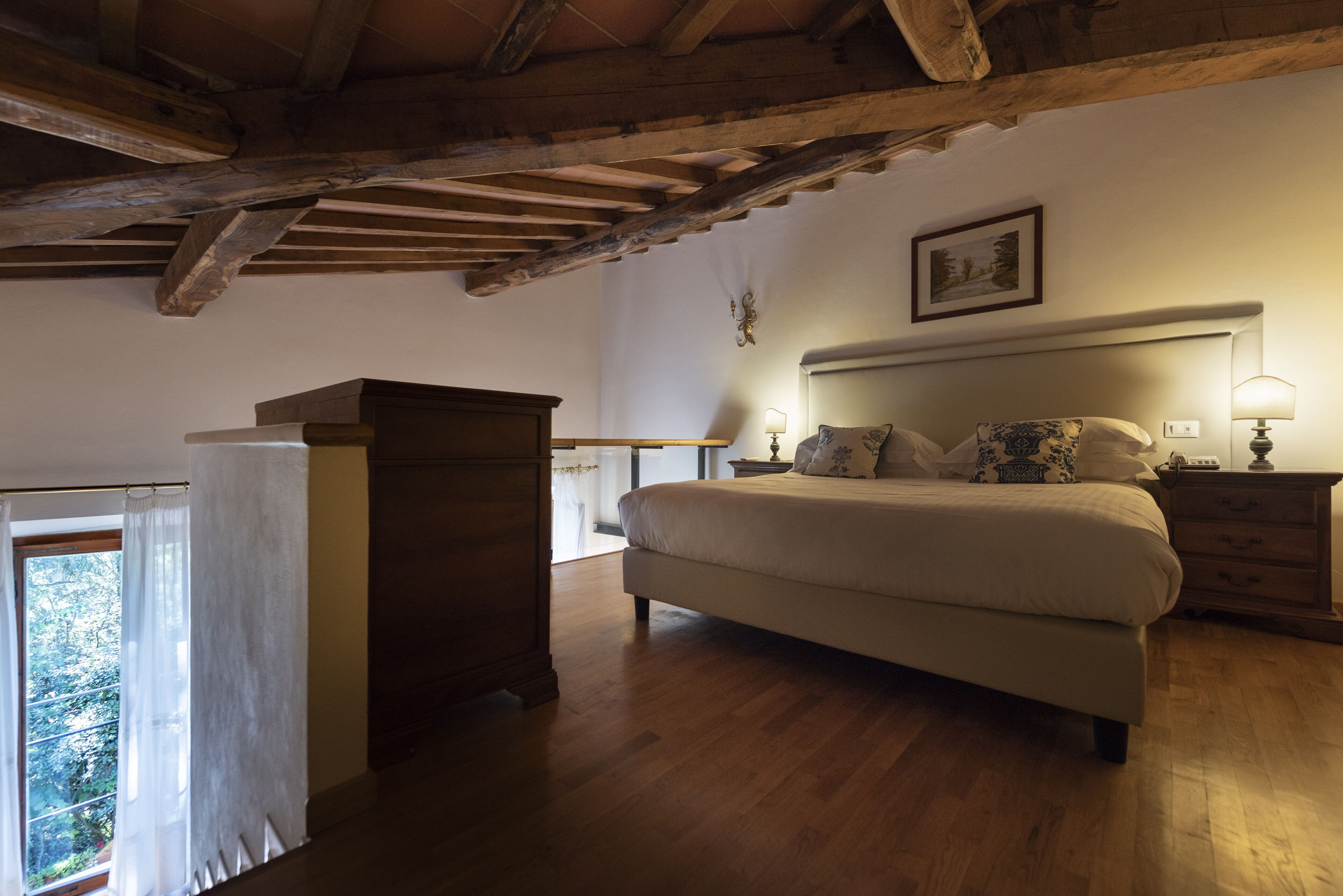 Ein Schlafzimmer mit einem großen Bett, Holzboden, einem Holztisch, einem Fenster mit Vorhängen und einem gerahmten Bild an der Wand.