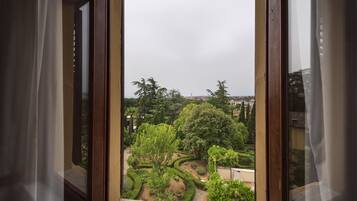 Vista a través de una ventana con cortinas, mostrando un jardín ajardinado con varios árboles y arbustos.