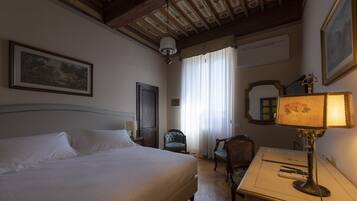 Quarto de hotel com uma cama grande, uma escrivaninha com abajur, uma cadeira e uma janela com cortinas.