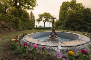 Una fontana circolare in pietra con una statua al centro, circondata da una varietà di fiori colorati e alberi in un giardino.