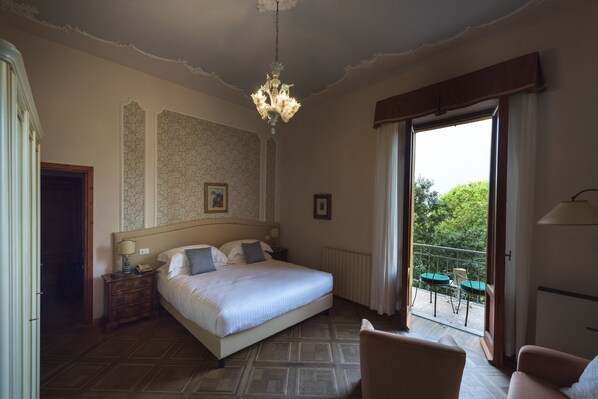 A bedroom with a large bed, a chandelier, a view of the outdoors through a balcony, and a wooden floor.