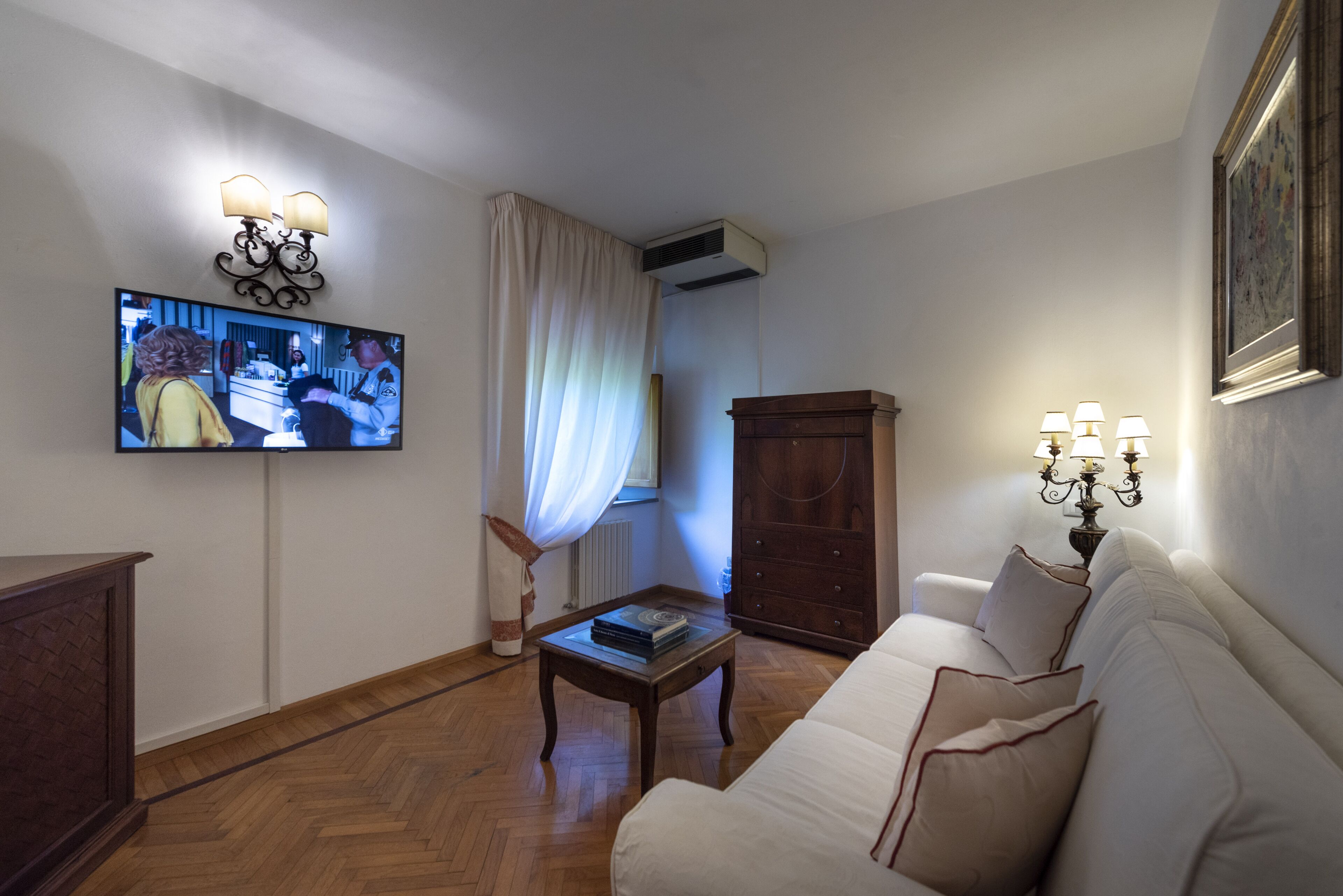Ein Wohnzimmer mit Fernseher, Couchtisch aus Holz, weißes Sofa, Holzschrank und Wandlampe.