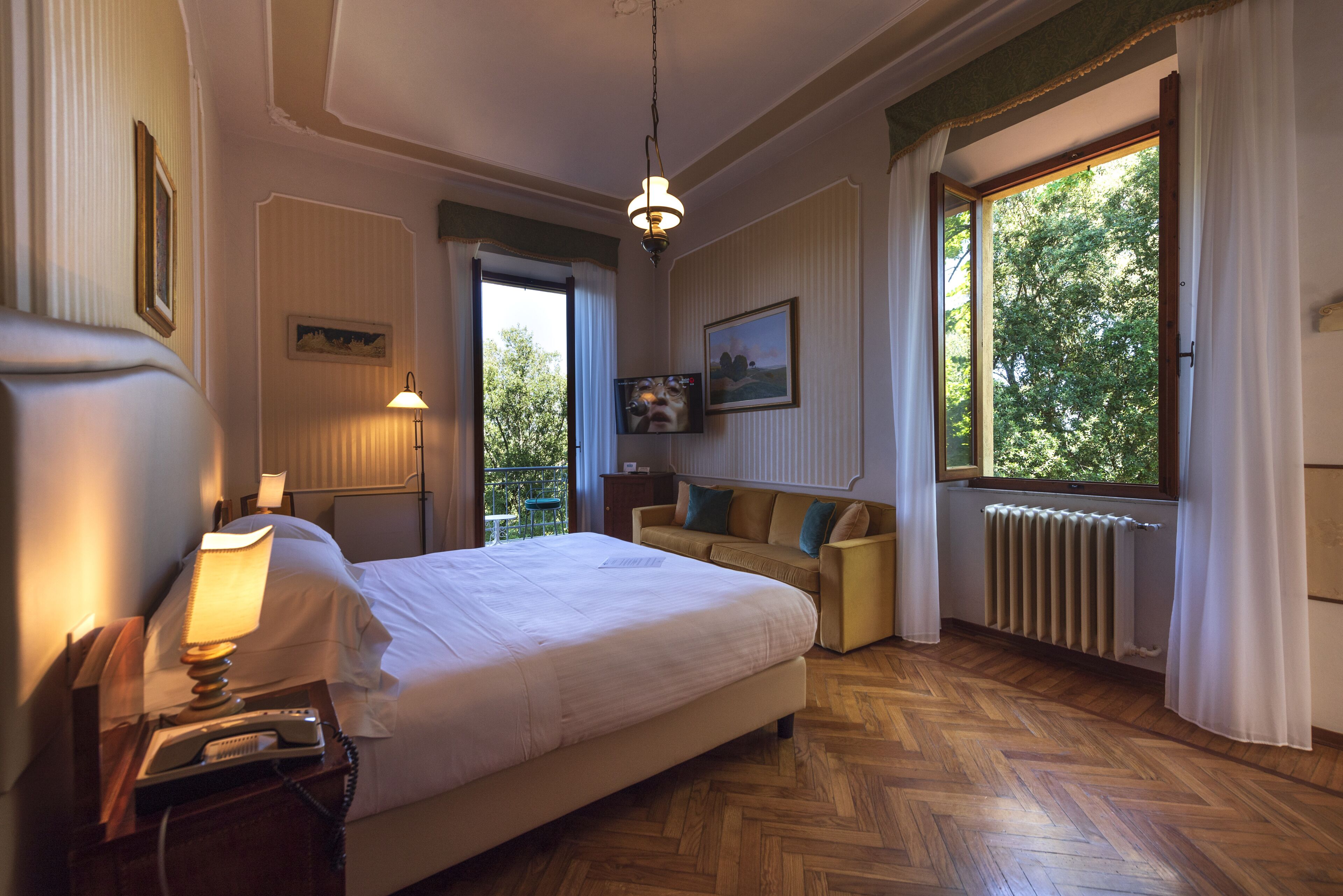 A hotel room with a large bed, a bedside lamp, a telephone, a sofa, and a window with a view of trees.