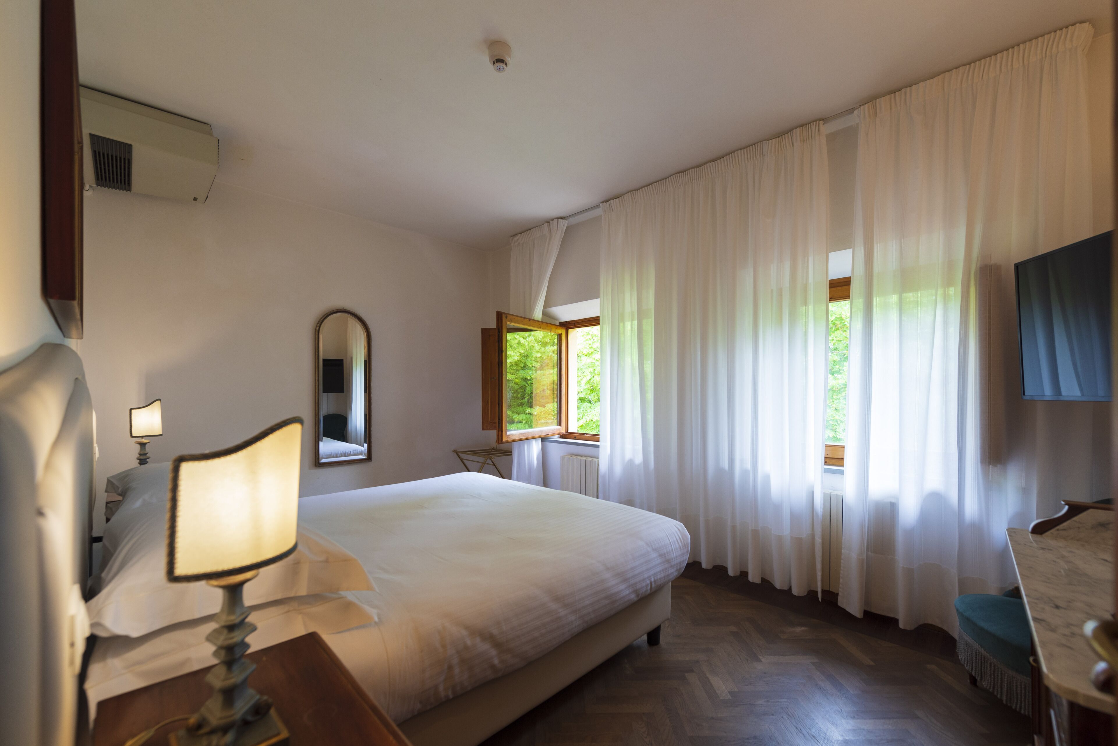 A hotel room with a large bed, a desk with a chair, a window with curtains, and a television on the wall.