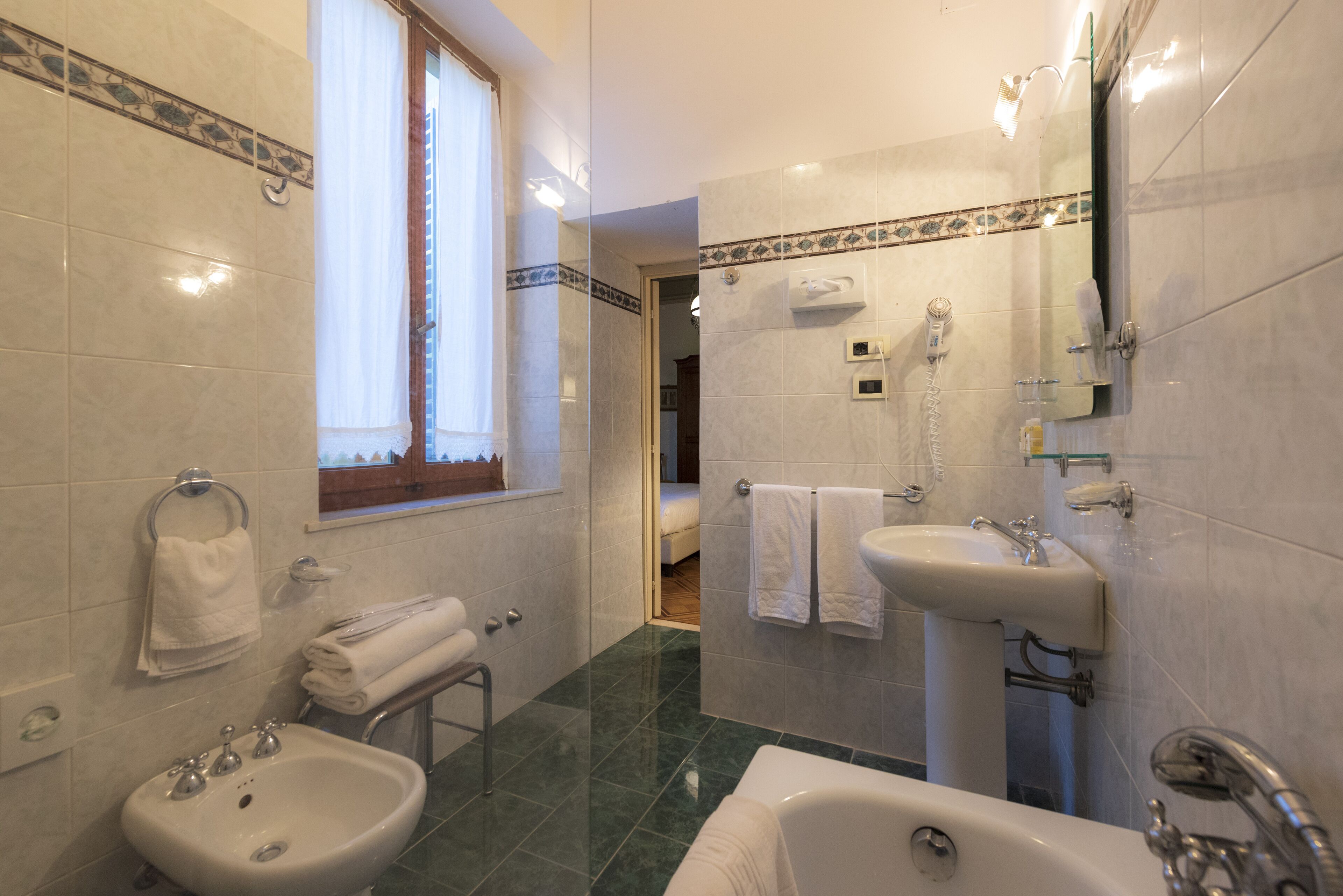 Un bagno con lavandino, vasca e doccia. Si nota una finestra con tende, un porta asciugamani e uno specchio.