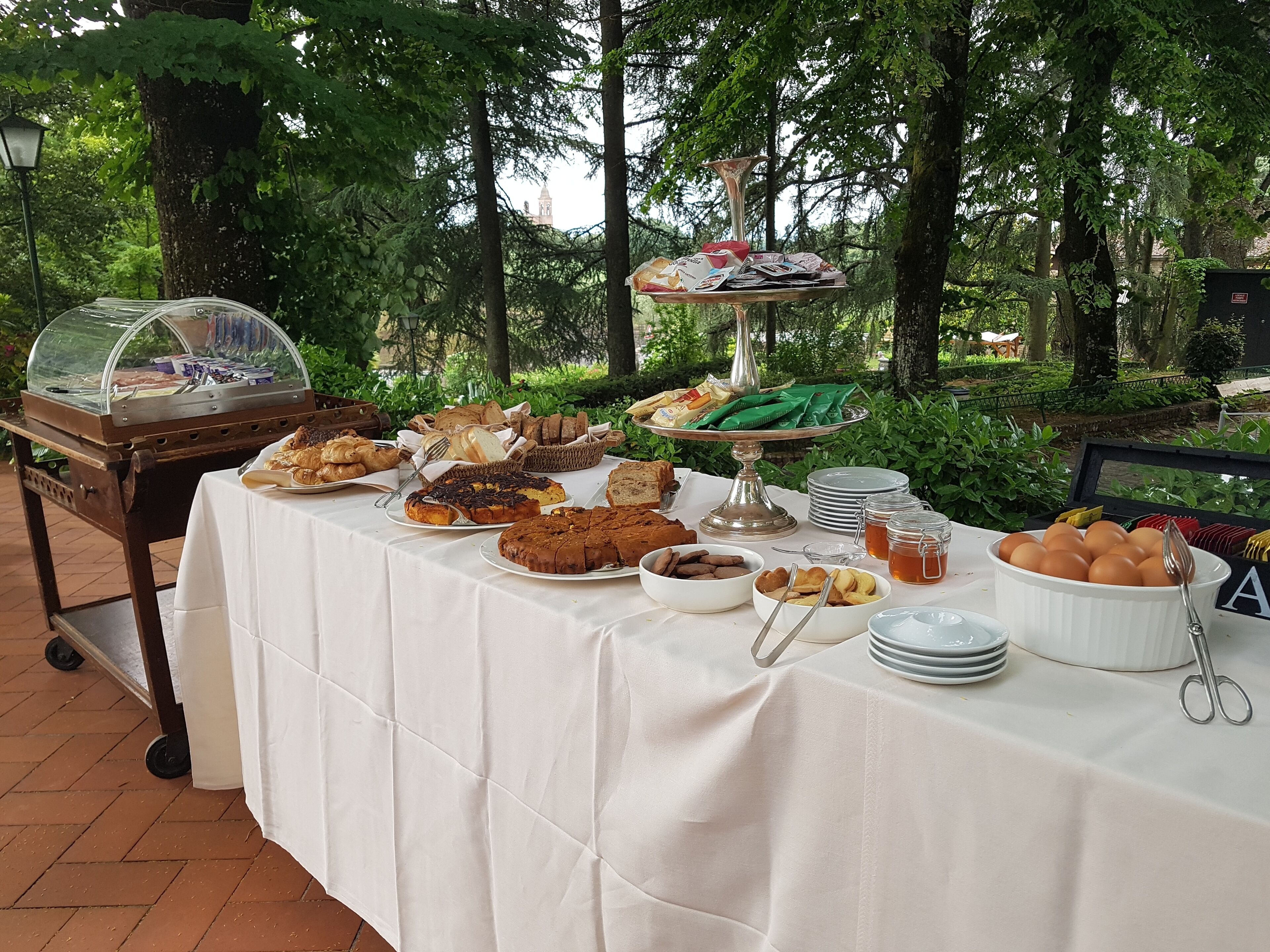 A buffet table with various food items and a dessert display, set outdoors with trees in the background.