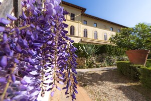 Un edificio con tetto in tegole e finestre ad arco, circondato da un vialetto sterrato e vegetazione.