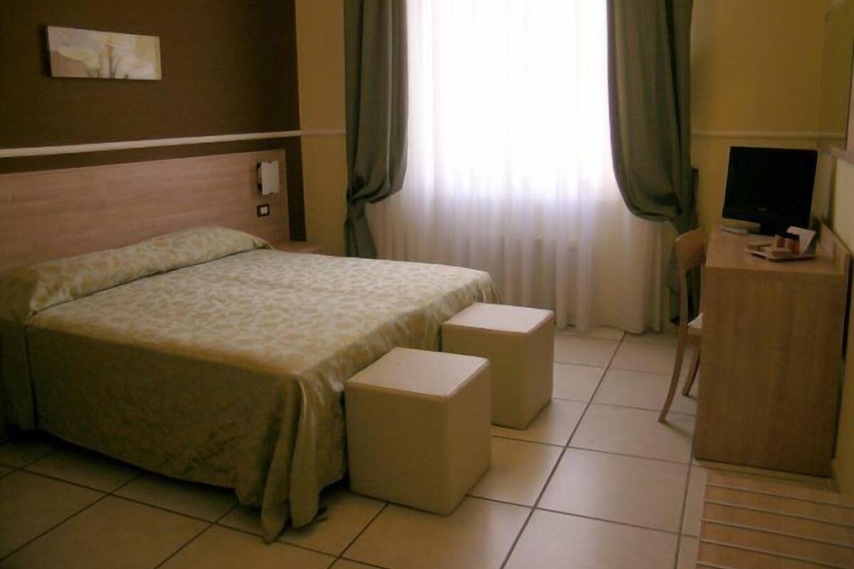 A hotel room with a bed, two bedside tables, a desk with a chair, a television, and a window with curtains.