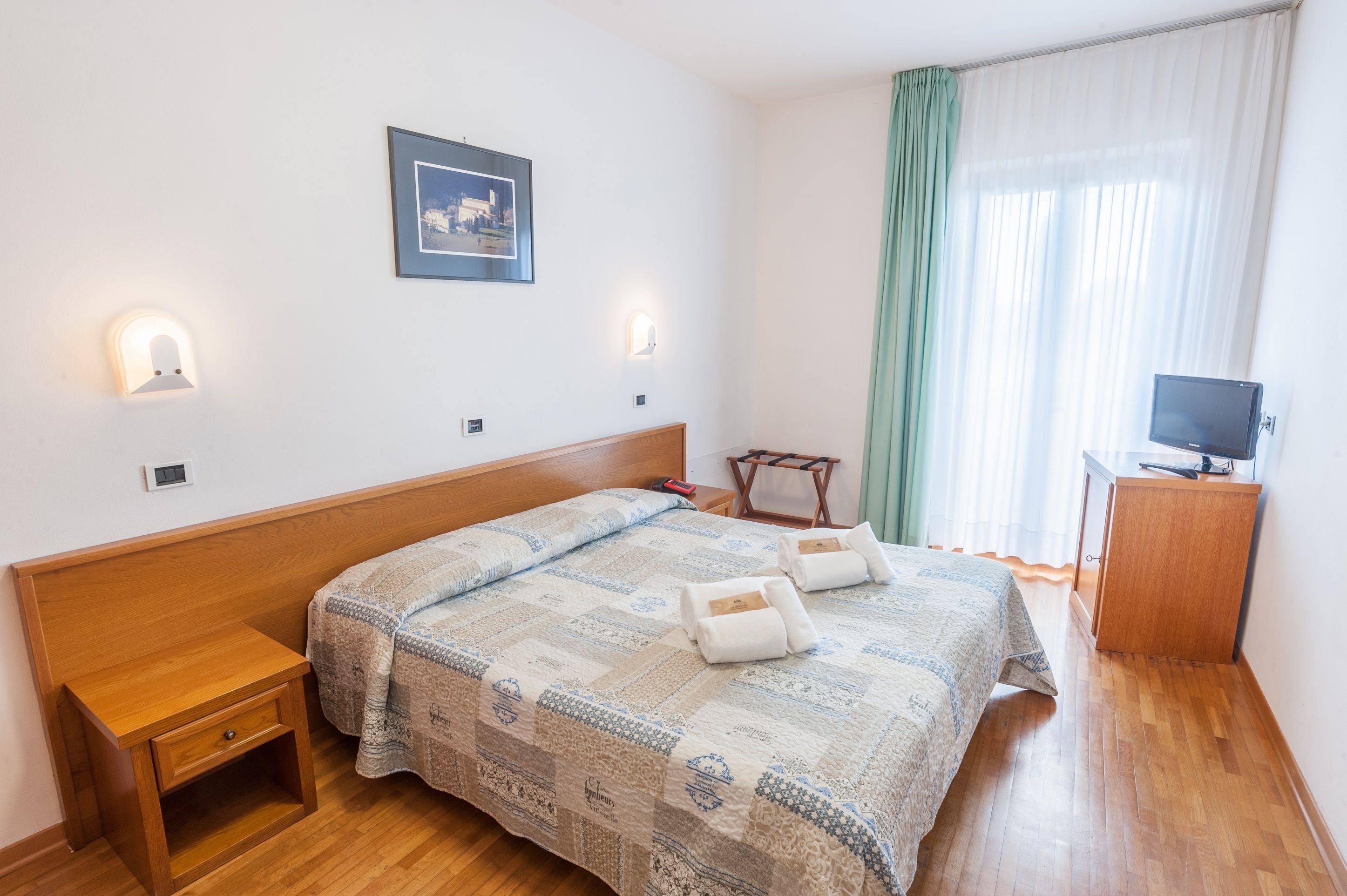 A neatly arranged bedroom with a wooden bed, bedside tables, a small desk, a television, and a framed picture on the wall.