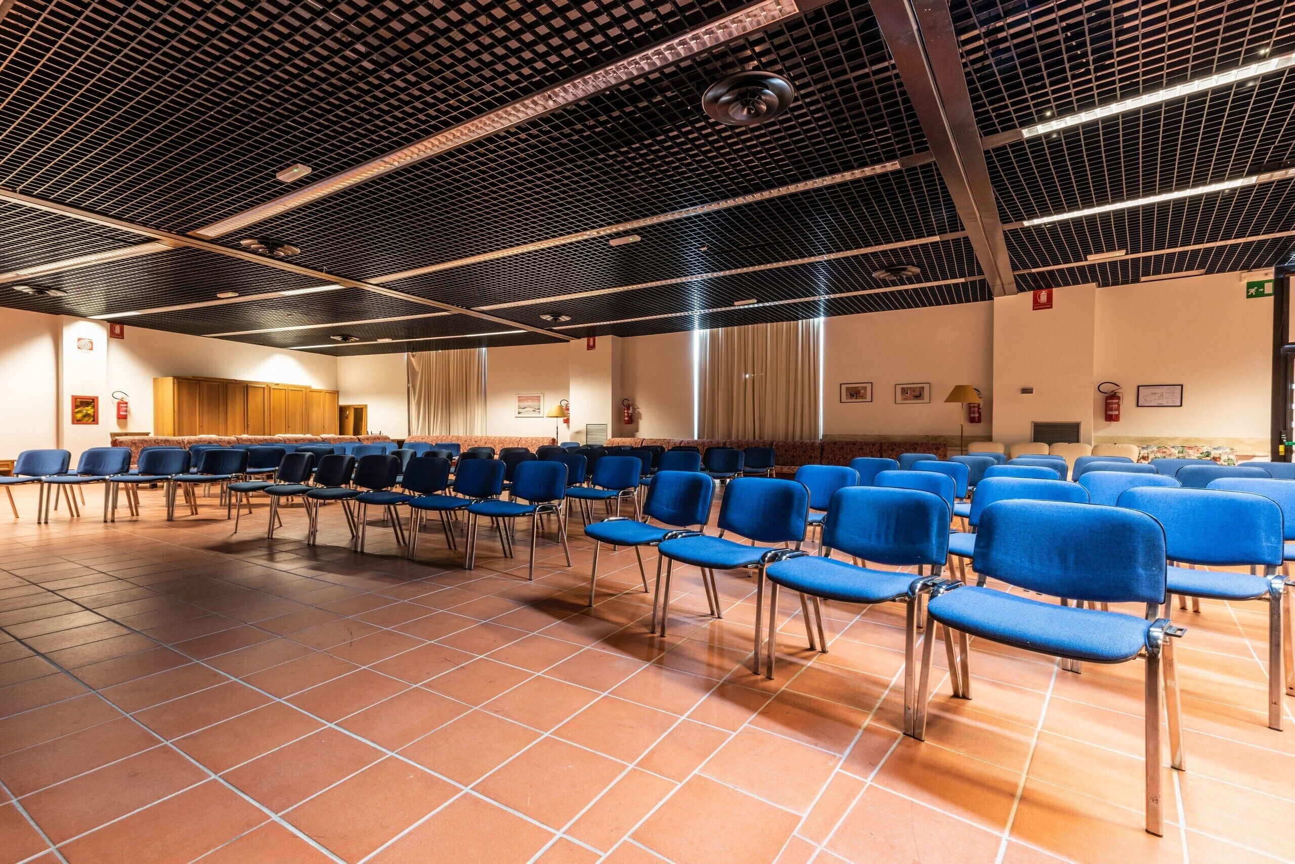 A conference room with rows of blue chairs, a patterned ceiling, and tiled flooring.