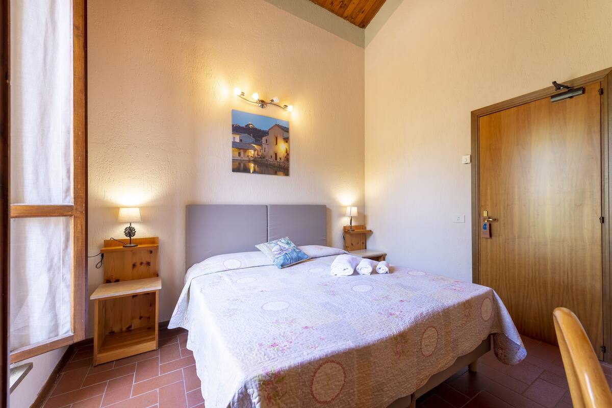Un dormitorio con cama, lámparas de noche, un pequeño estante de madera, una mesita de noche, una puerta de madera y un cuadro colgado en la pared.