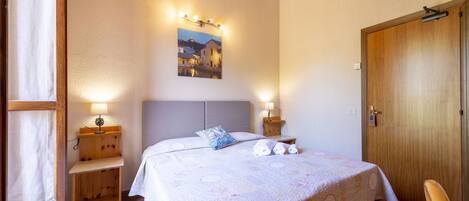 Un dormitorio con cama, lámparas de noche, una mesita de noche de madera, una puerta y un cuadro en la pared.