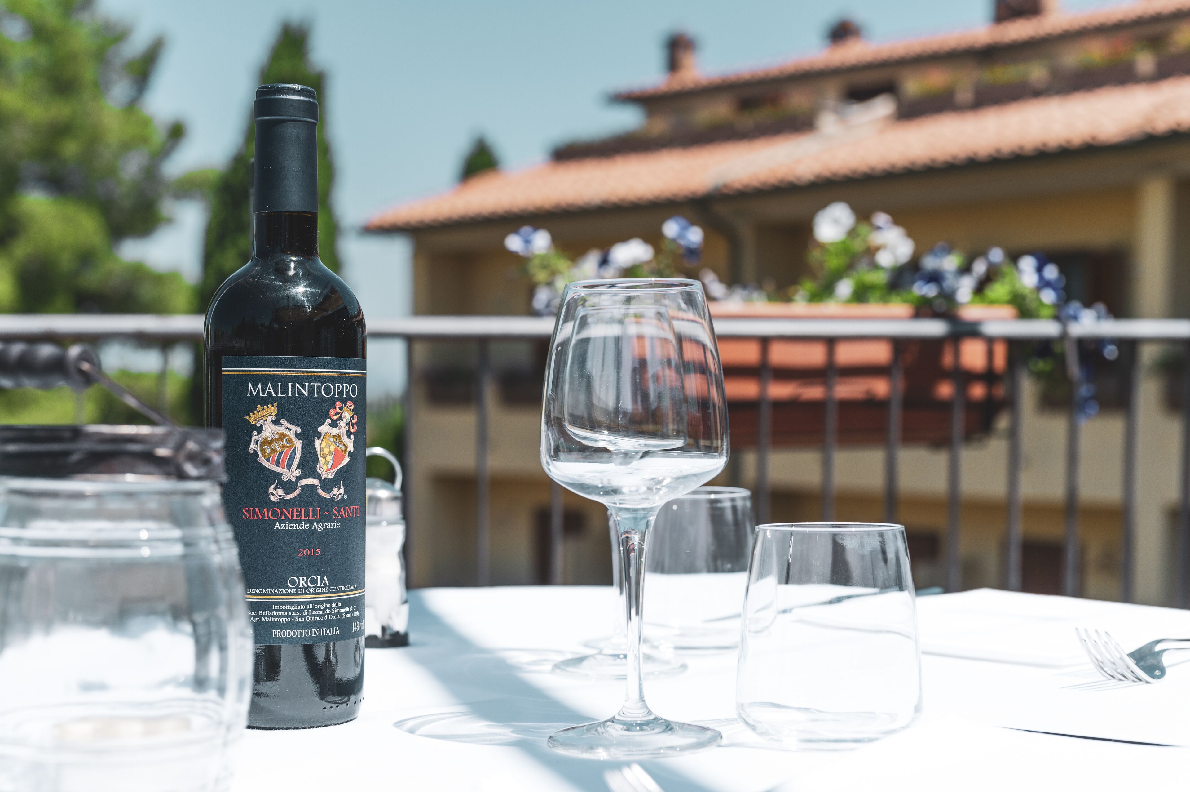 Une bouteille de vin Malintoppo et des verres à vin vides sont posés sur une table, avec un bâtiment visible en arrière-plan.