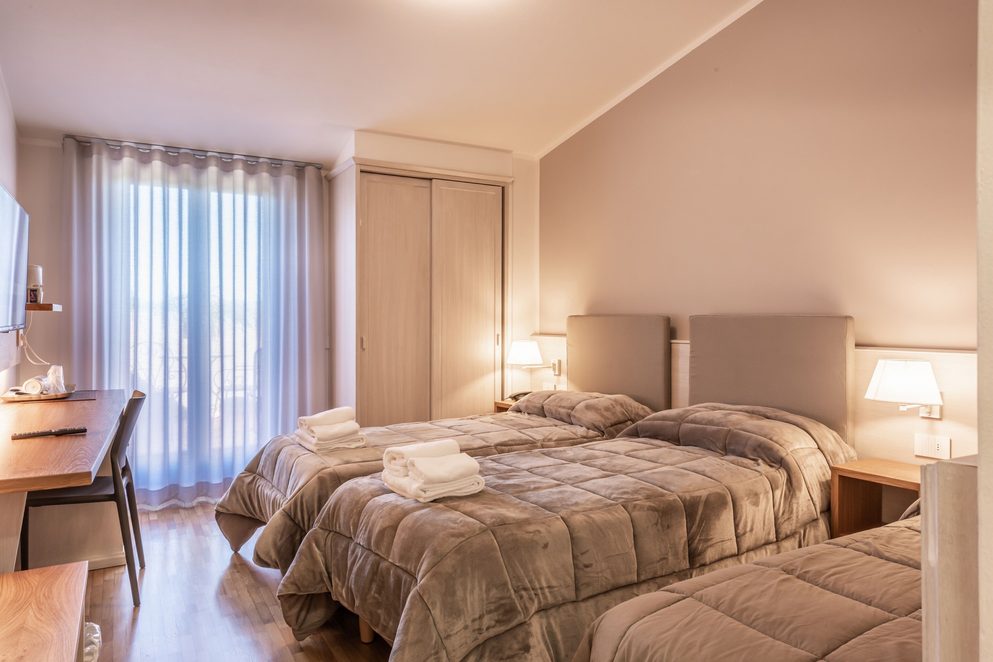 A hotel room with two beds, a desk, and a chair. There are lamps on the bedside tables and a window with curtains.
