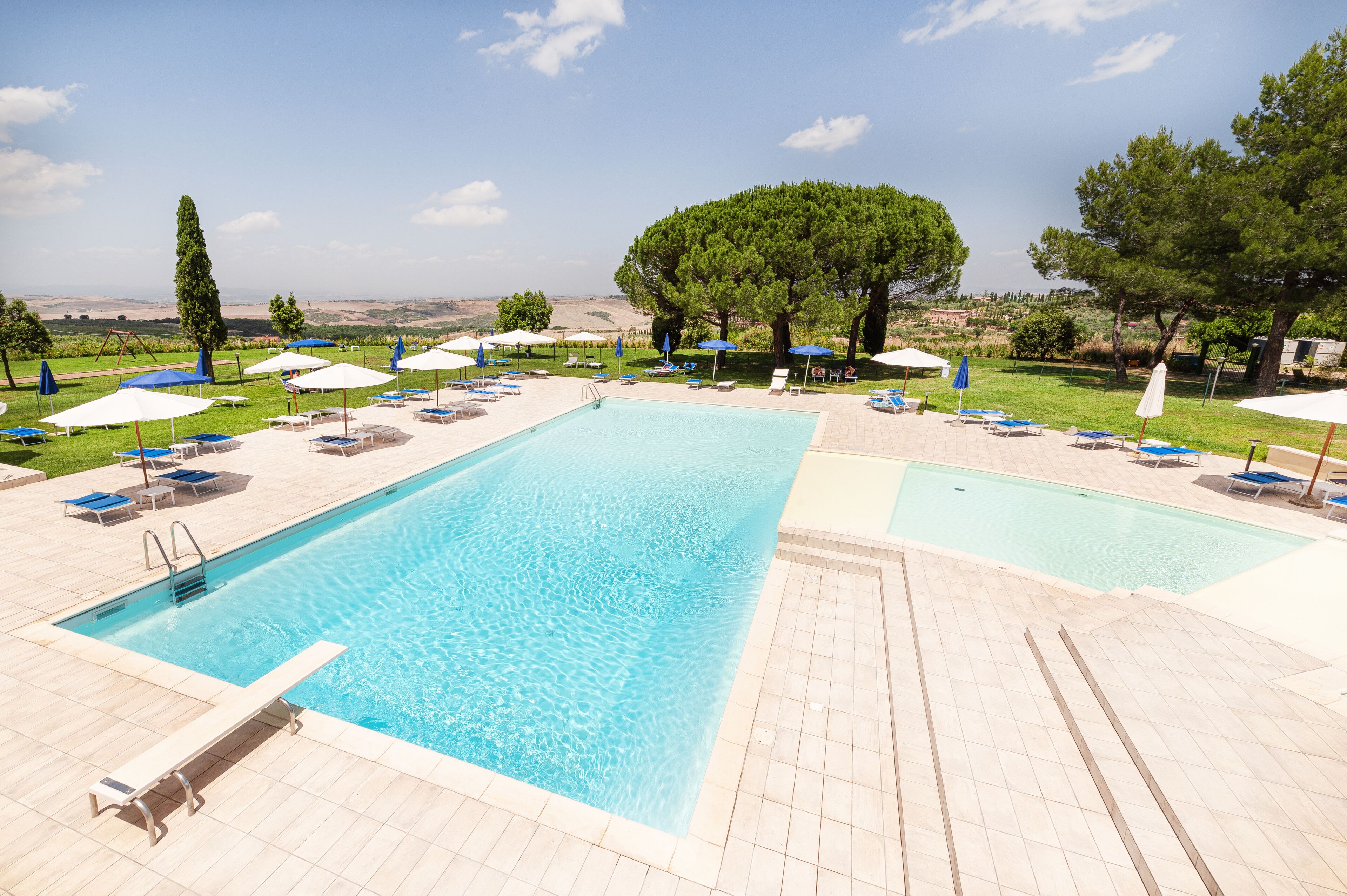 A swimming pool with sun loungers and umbrellas, surrounded by trees and a view of the countryside.